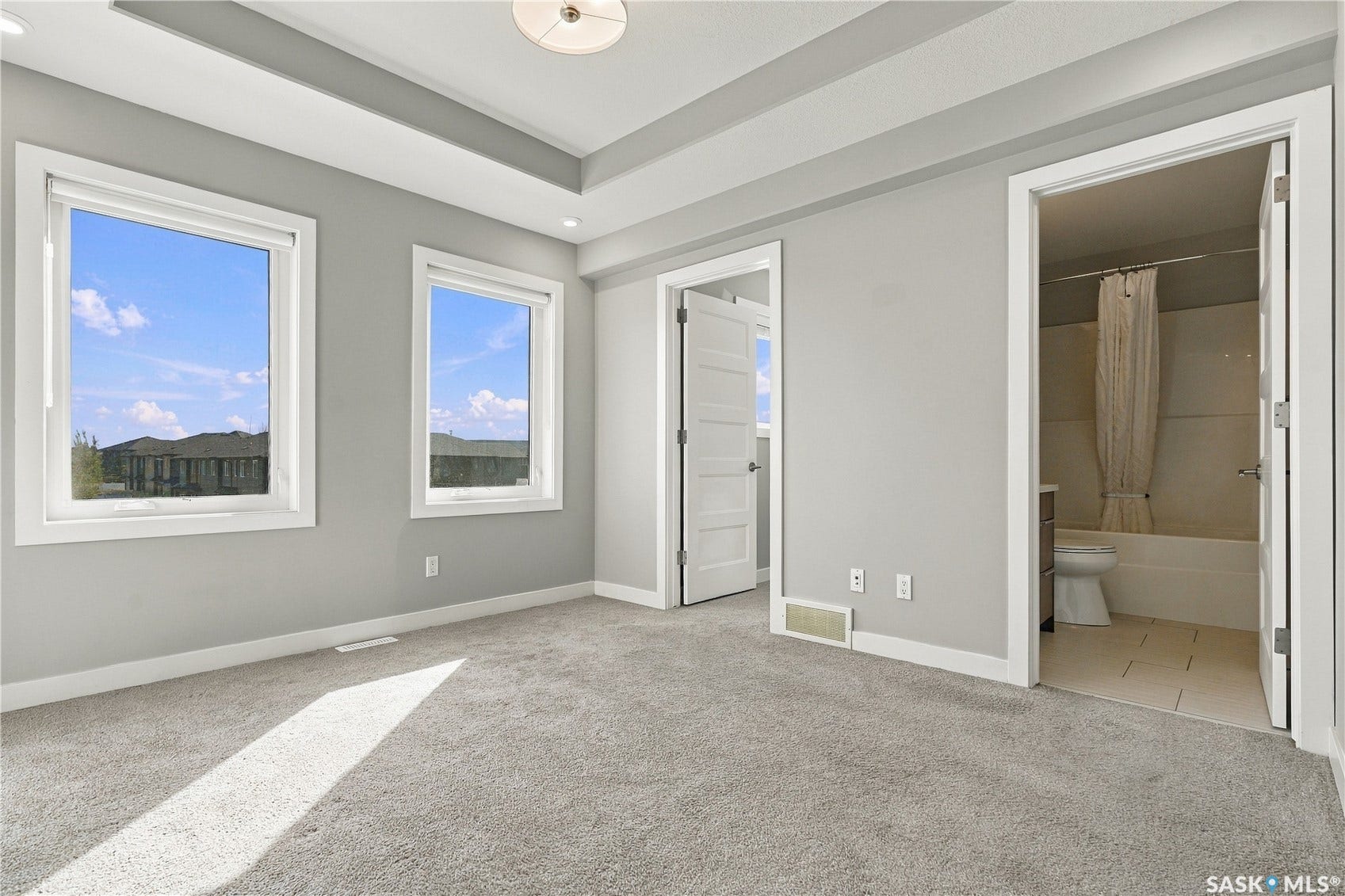 Recessed ceiling with an overhead light fixture, two windows with white trim, light grey carpeting, and an adjacent bathroom with a bathtub