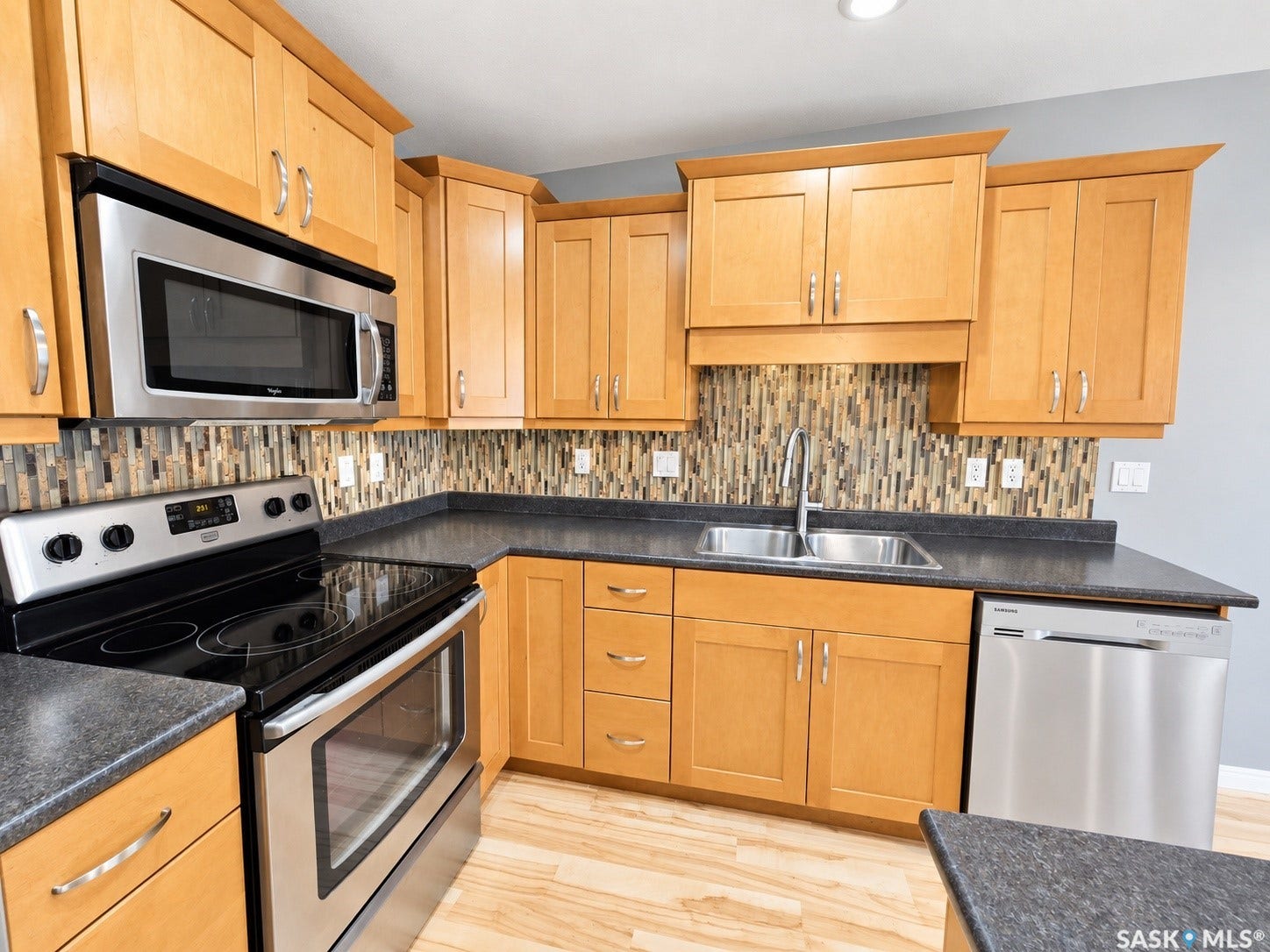 Wood-finish flooring, stainless steel appliances, dual basin sink, light wood cabinetry, and a textured tile backsplash