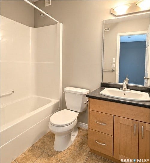 Tile flooring, light wood-finish vanity, integrated sink, dark countertop, and a white tub/shower combination