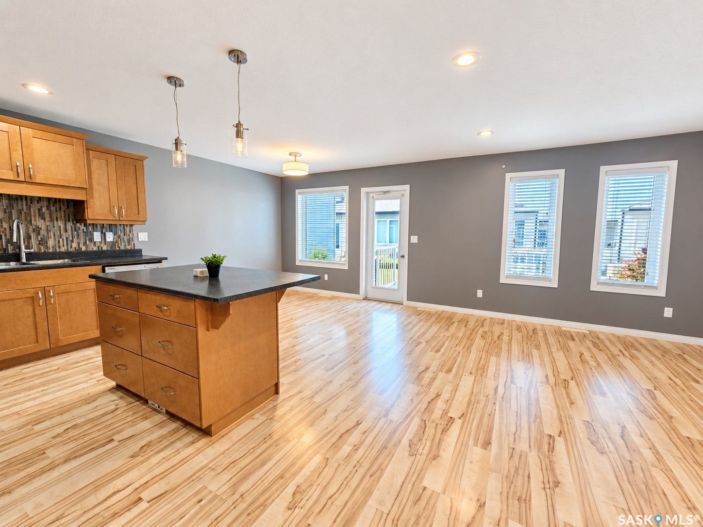Wood-finish flooring throughout, featuring a kitchen island with dark countertop, light wood cabinetry, and a patterned tile backsplash