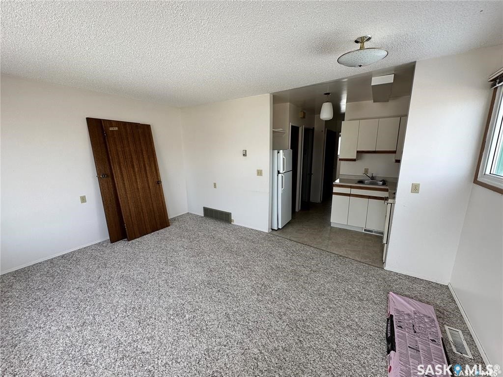 White walls with a textured ceiling, grey carpet, and a kitchen area featuring white cabinetry and a white refrigerator