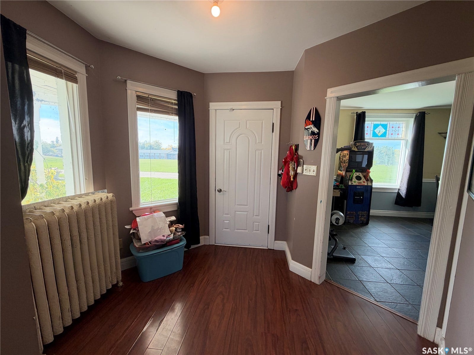 Wood-finish flooring, two windows with blinds, a white paneled door, and a traditional radiator