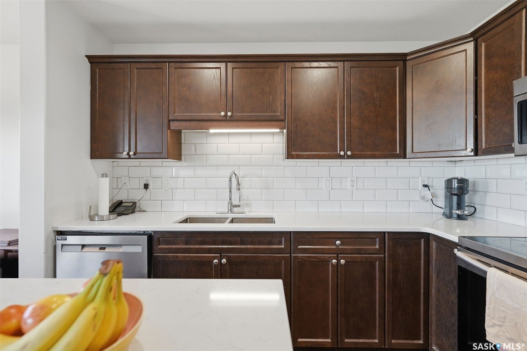 Kitchen featuring wood-finish cabinetry, white subway tile backsplash, white countertops, stainless steel undermount sink, and stainless steel dishwasher