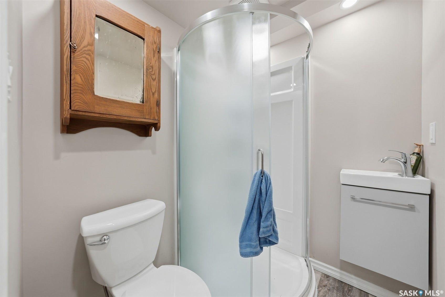 Frosted glass shower enclosure, wall-mounted vanity with integrated sink, chrome faucet, white toilet, and wood-finish wall cabinet