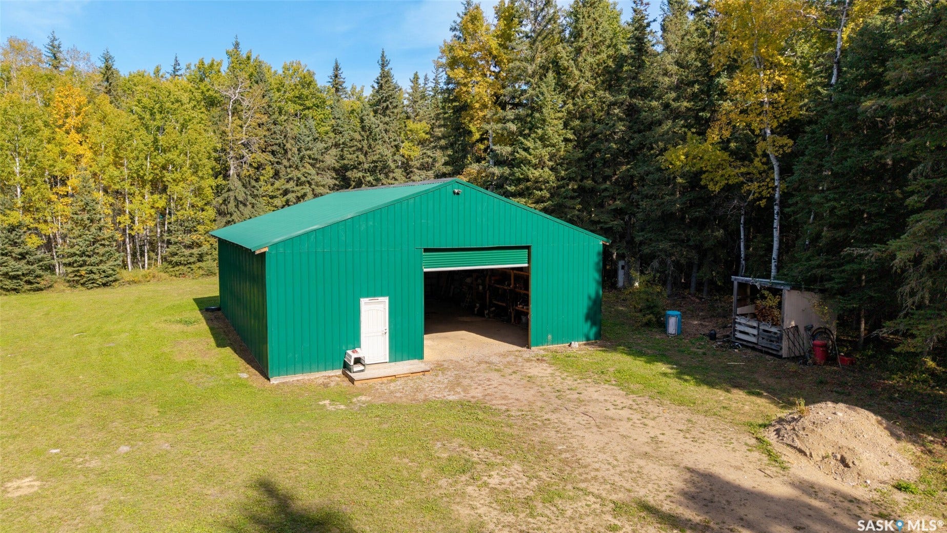 Green metal-sided outbuilding with an overhead door, adjacent to an expanse of natural landscaping and mature trees