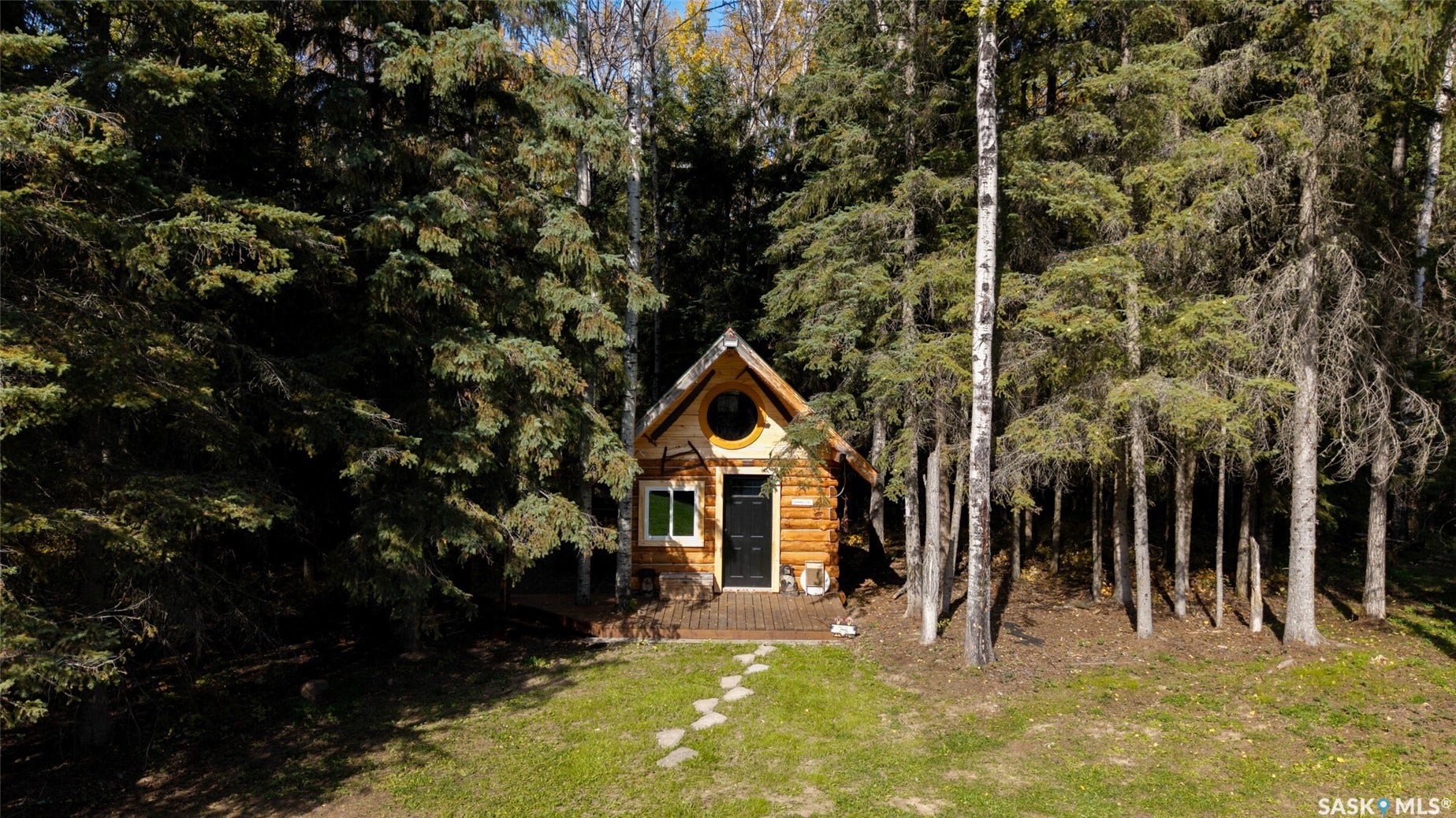 Log cabin construction with a circular window, surrounded by mature trees, featuring a wooden deck and a stone path