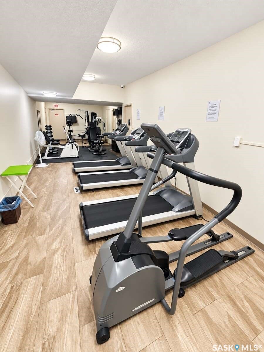 State-of-the-art fitness facility featuring multiple treadmills and stationary bikes with gray upholstery and black frames, wood-finish flooring throughout, recessed and pendant lighting fixtures, and ample square footage for comprehensive workout space