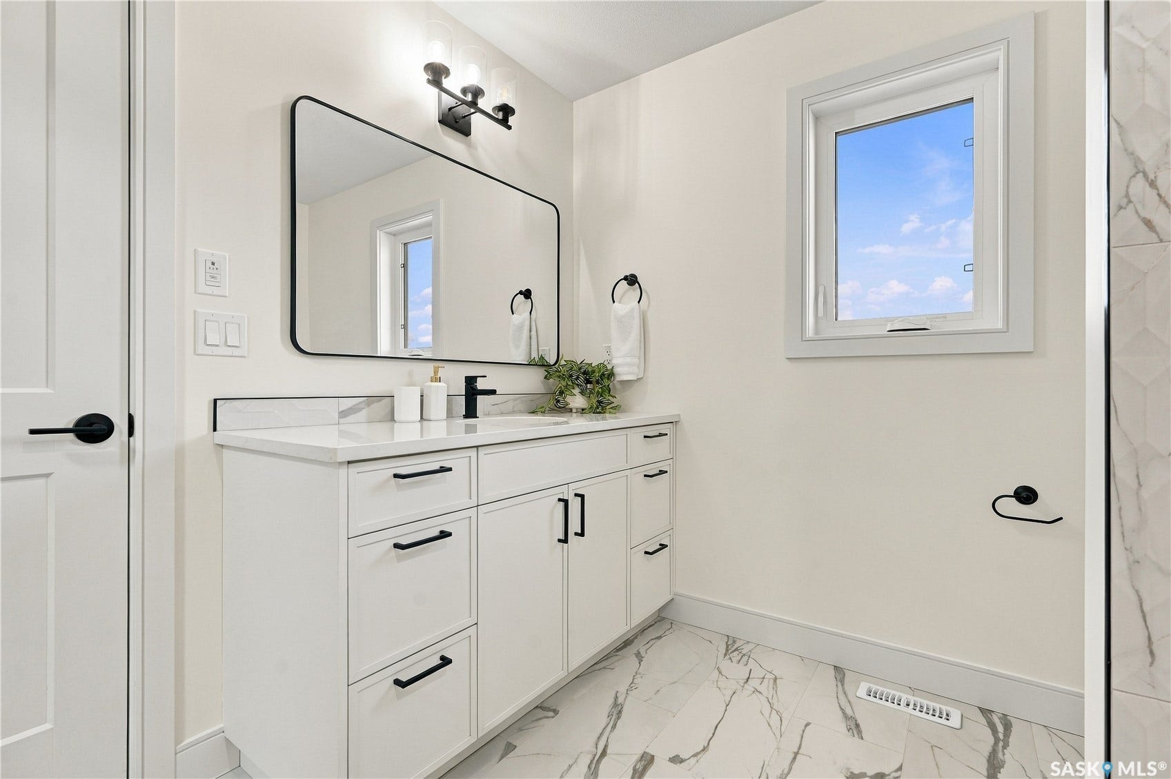 White vanity with integrated sink, black hardware, framed mirror, wall-mounted lighting, and marble-patterned flooring