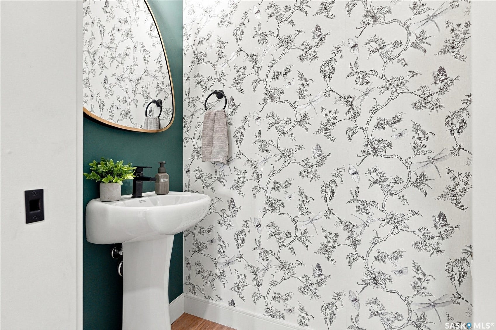 Pedestal sink with a black finish faucet, oval mirror with a gold-tone frame, and decorative wallpaper