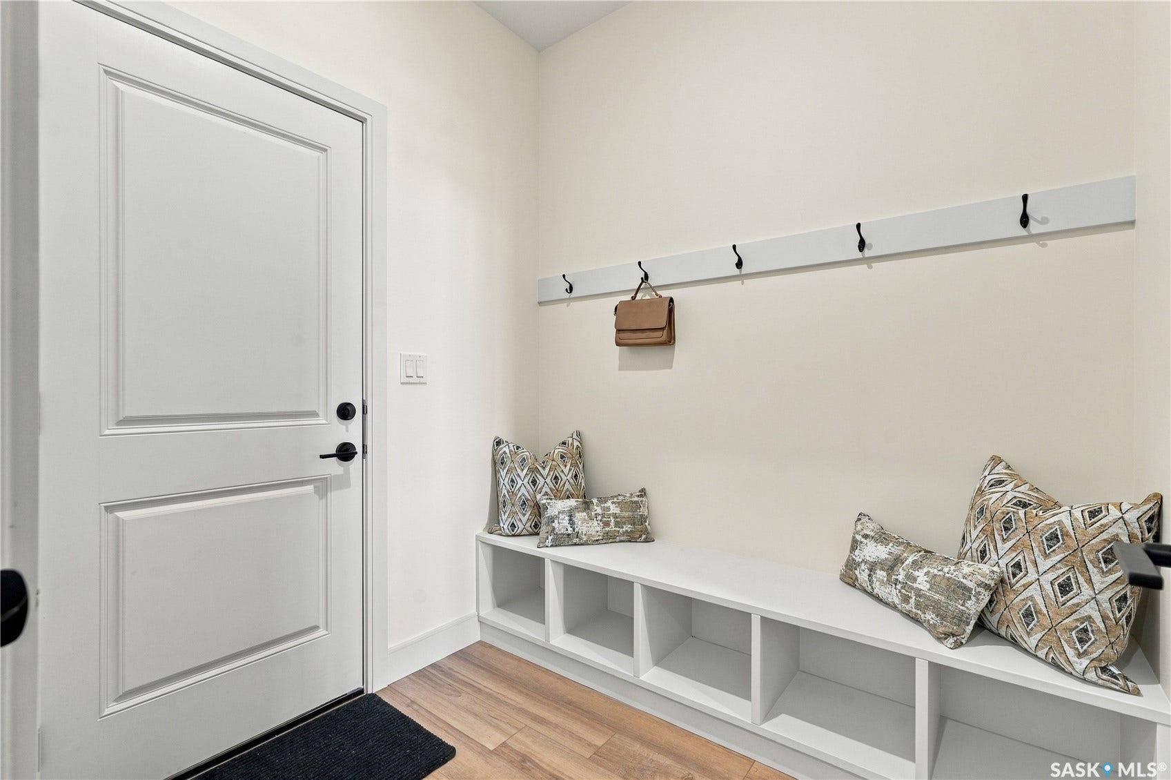 Built-in storage bench with open cubbies, wood-finish flooring, white paneled door, and wall-mounted coat hooks