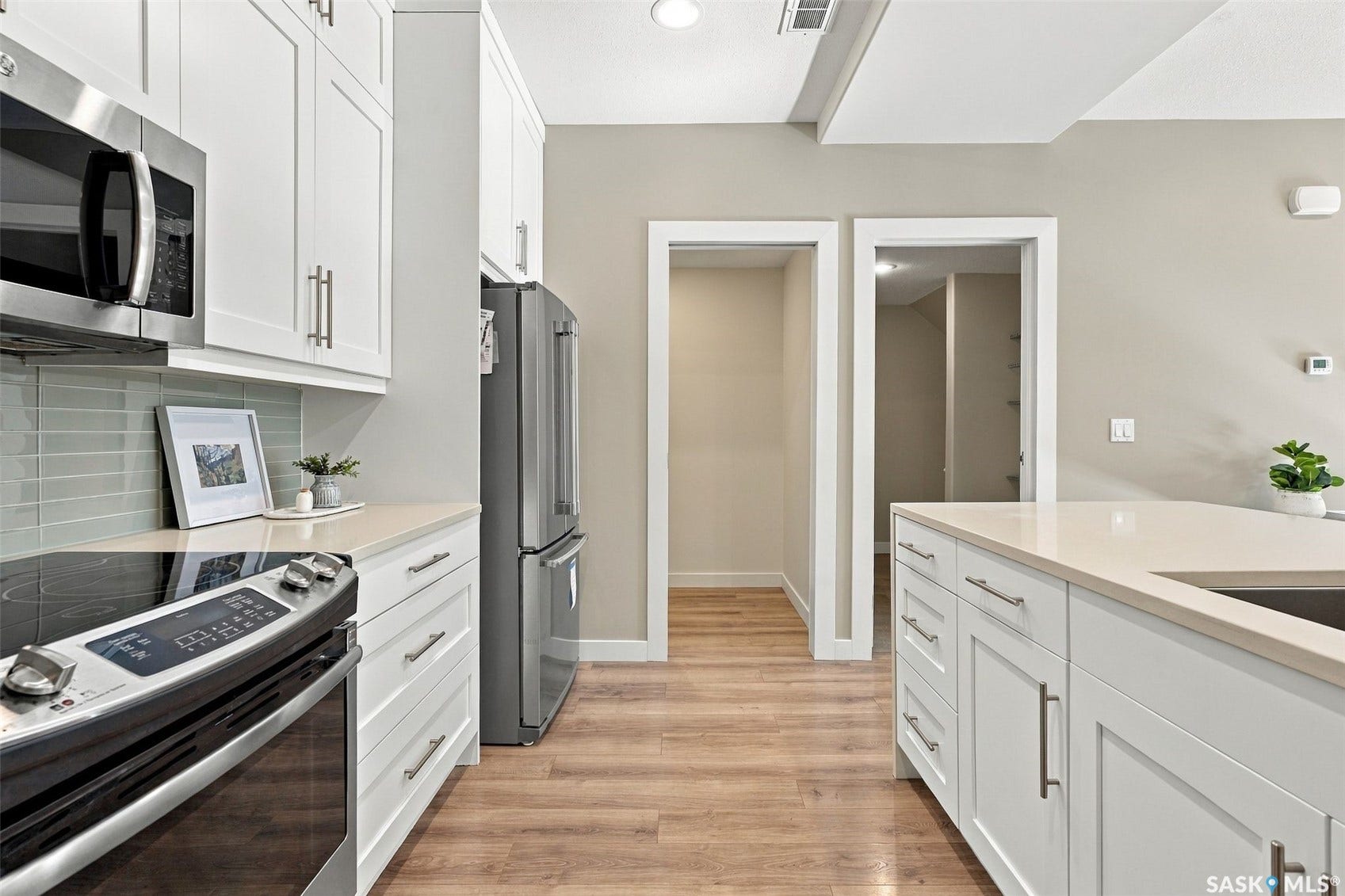 White shaker-style cabinetry with brushed nickel hardware, stainless steel appliances, light-toned countertops, horizontal tile backsplash, and wood-finish flooring