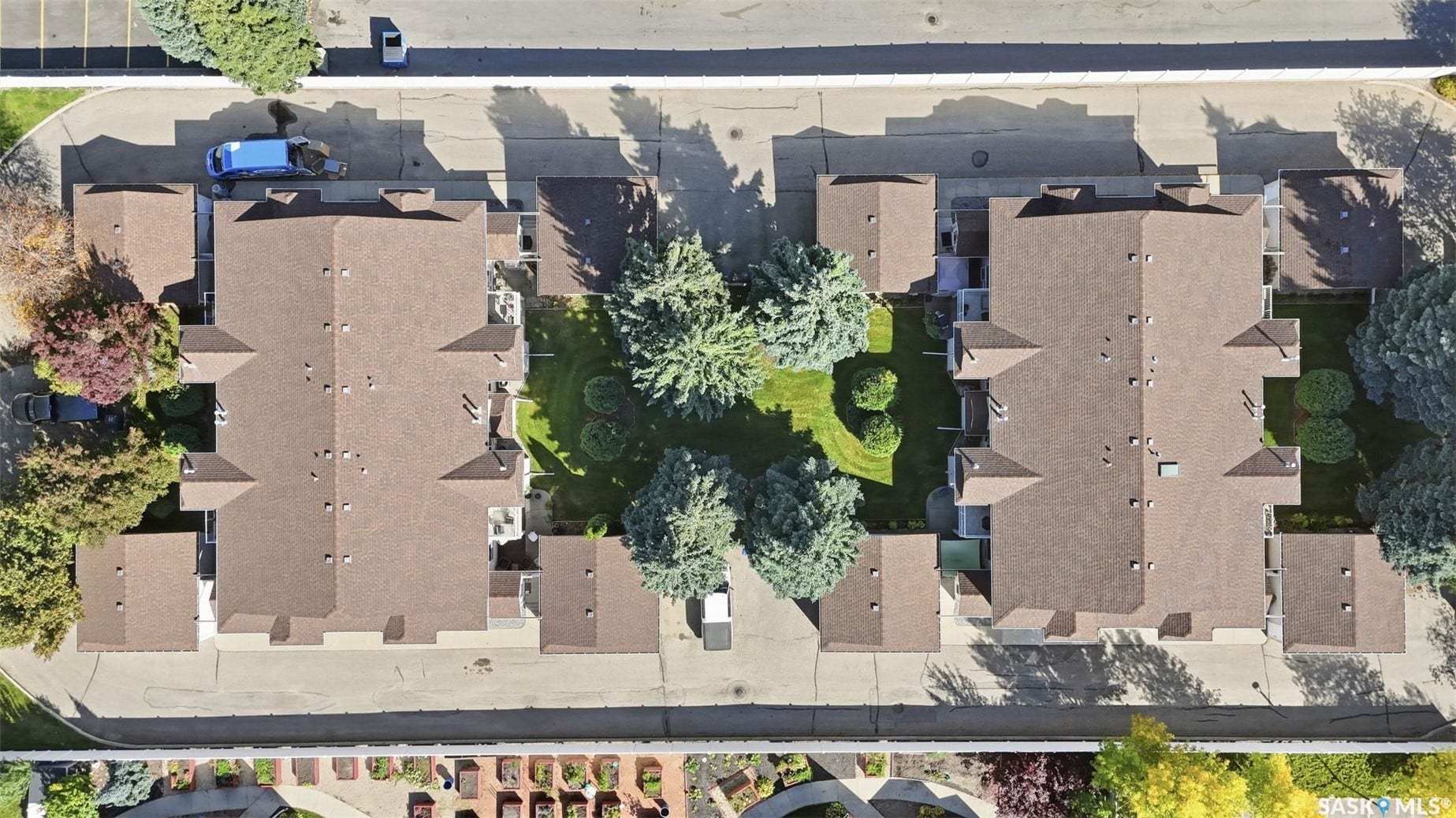 Two multi-unit residential buildings with brown shingle roofing, surrounded by mature trees and paved parking areas
