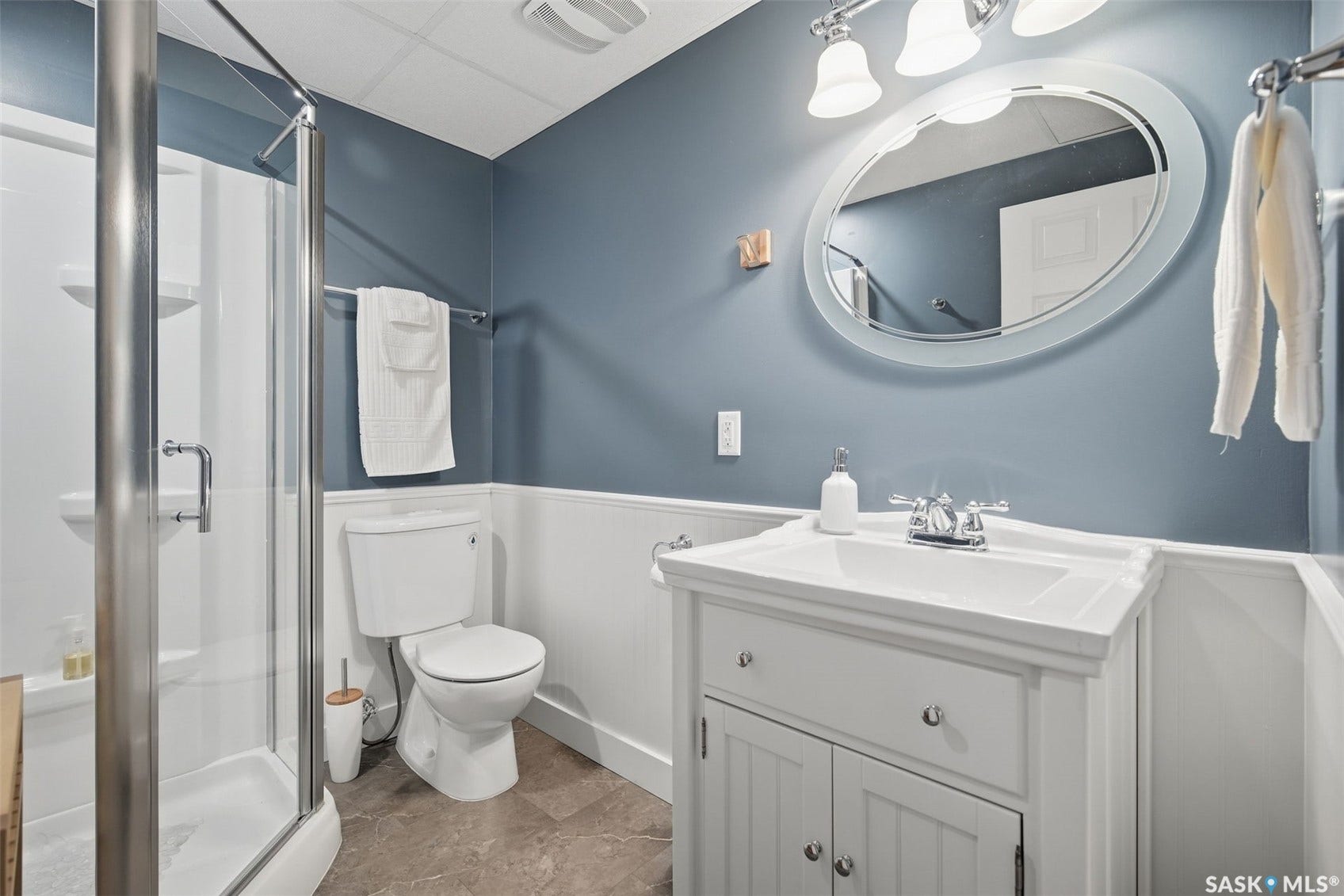White vanity with integrated sink, chrome faucet, oval wall mirror, glass-enclosed shower, and tile flooring