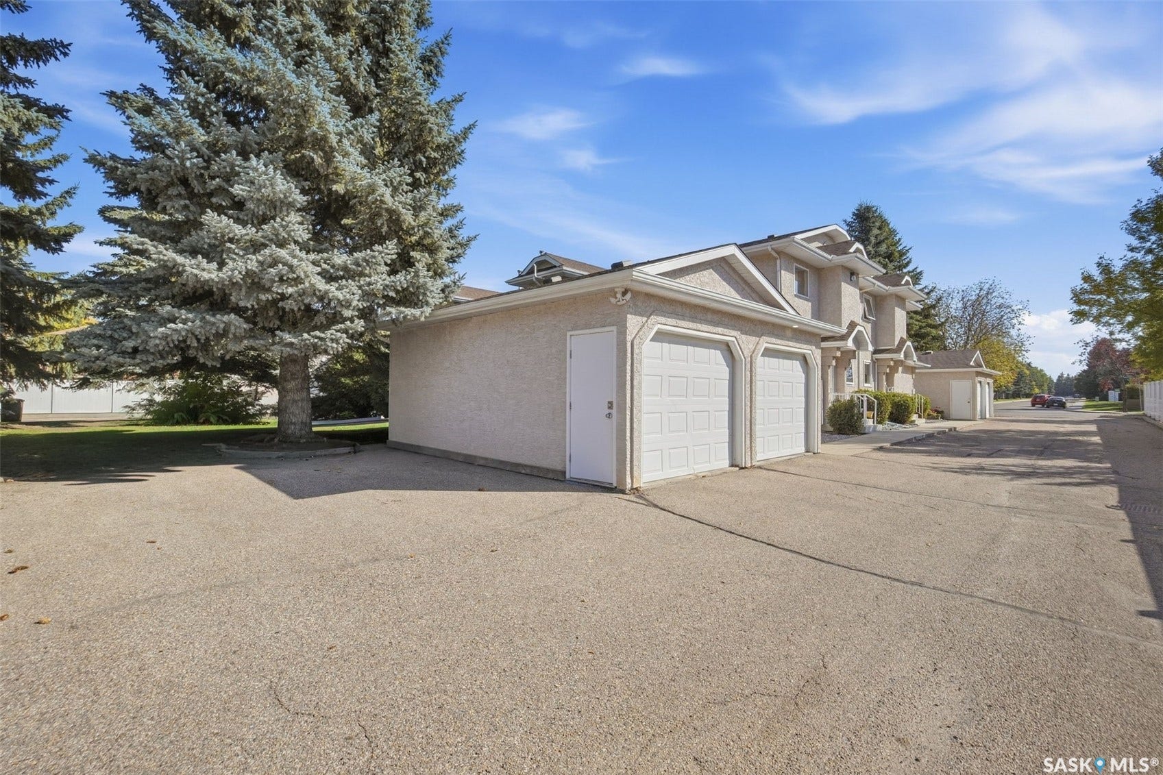 Stucco exterior with gabled roof lines, featuring a two-car garage with panel doors, mature evergreen trees, and a paved driveway