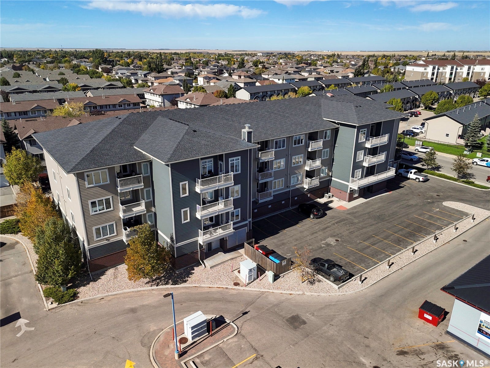 Multi-story building featuring balconies, dark gray shingled roof, light-colored siding, and a paved parking area