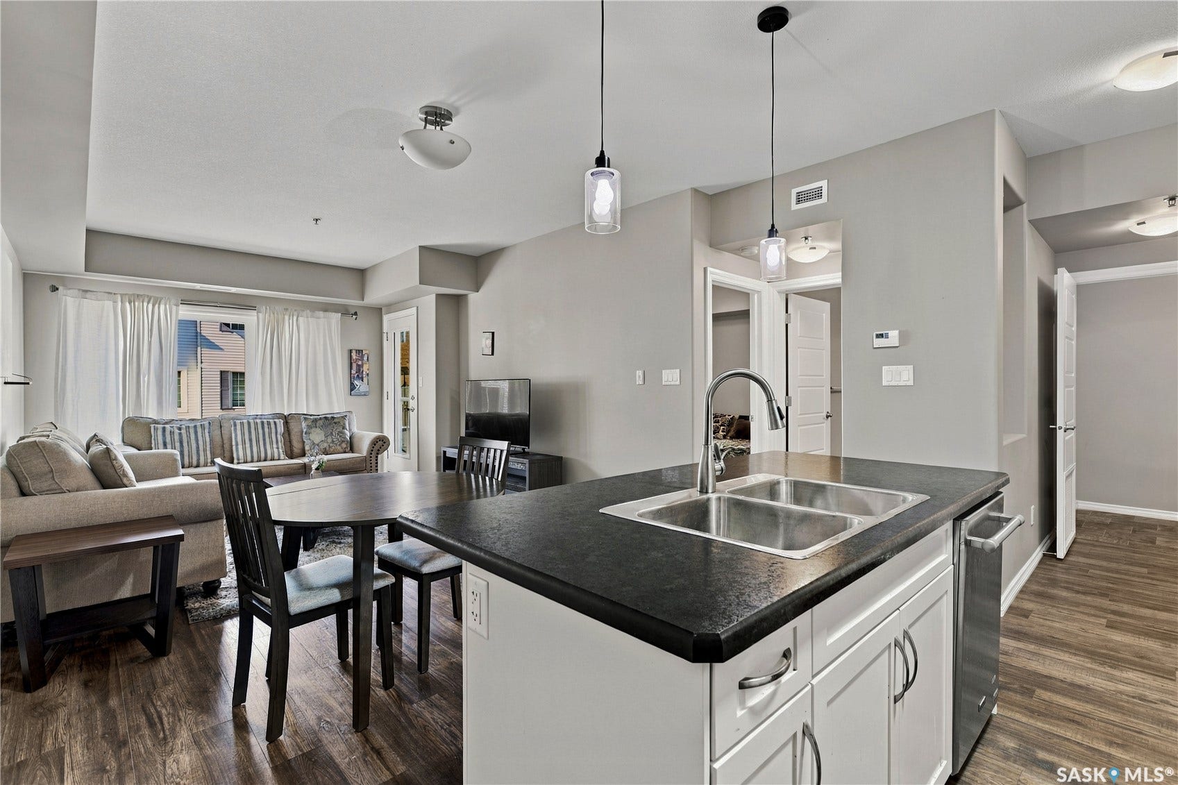 Open-concept kitchen and living area featuring a large island with black countertop and white cabinetry, stainless steel appliances, pendant lighting, wood-finish flooring throughout, and abundant natural light from multiple windows
