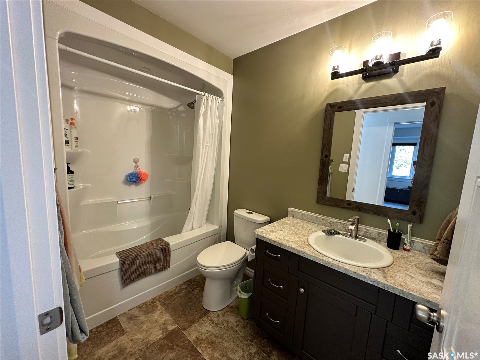 White tub-shower combination, dark wood-finish vanity with a light-toned countertop, framed mirror, and a three-light fixture