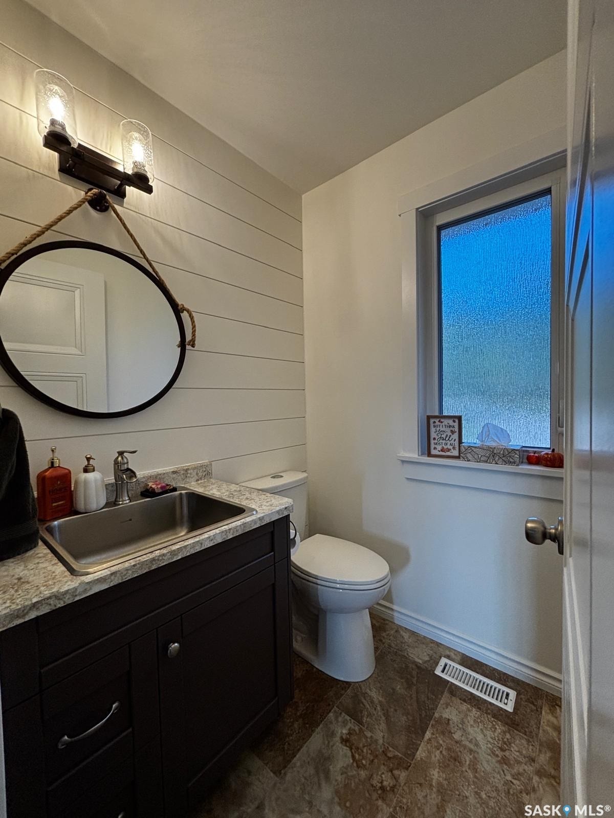 1/2 bathroom directly off front door with Shiplap accent wall, dark wood-finish vanity with stainless steel sink, patterned countertop, and neutral-toned flooring