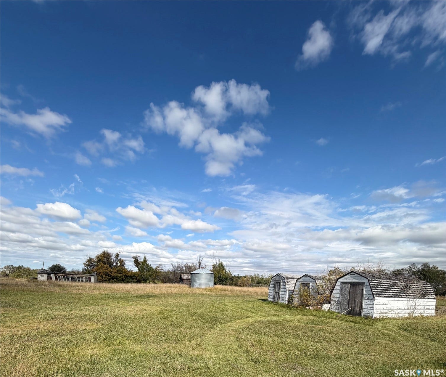 Expansive open field with scattered buildings, including a corrugated metal grain bin and multiple gambrel-roofed structures