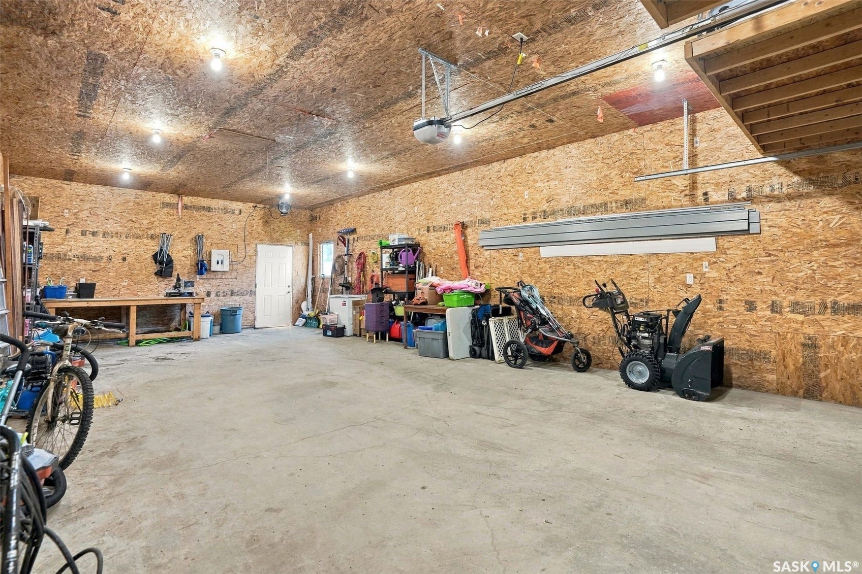 Spacious garage with concrete flooring, exposed oriented strand board walls and ceiling, multiple recessed ceiling lights, a workbench, and an overhead storage area