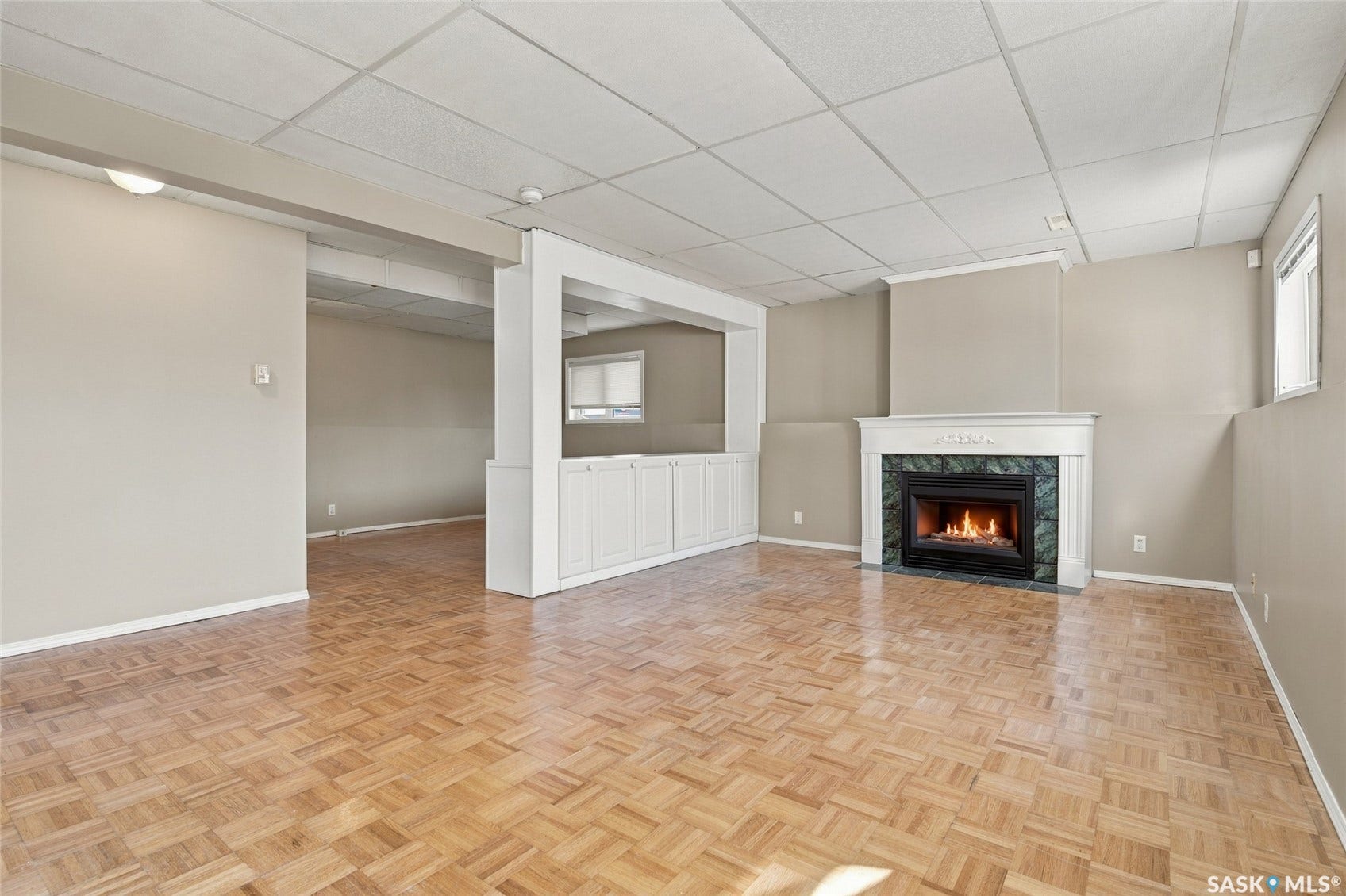 Parquet wood-finish flooring, white built-in cabinetry, white framed fireplace with stone surround, drop ceiling, and neutral painted walls