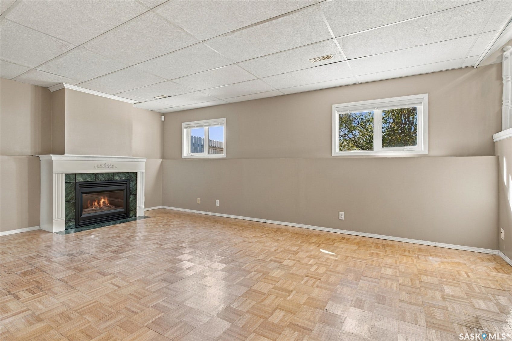 Wall-mounted fireplace with a white mantel and green surround, two windows, wood-finish flooring, and a drop ceiling