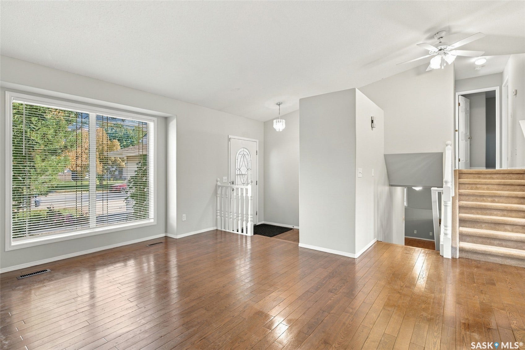 Hardwood-style flooring, large picture window, white-painted trim, white entry door with decorative glass insert, and a ceiling fan