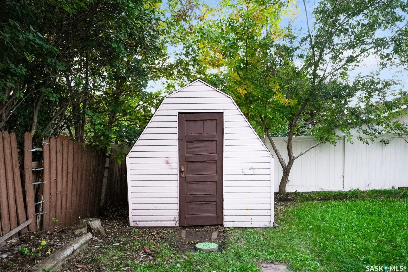 Charming backyard storage structure featuring white horizontal siding, arched roof design, and dark wood paneled door