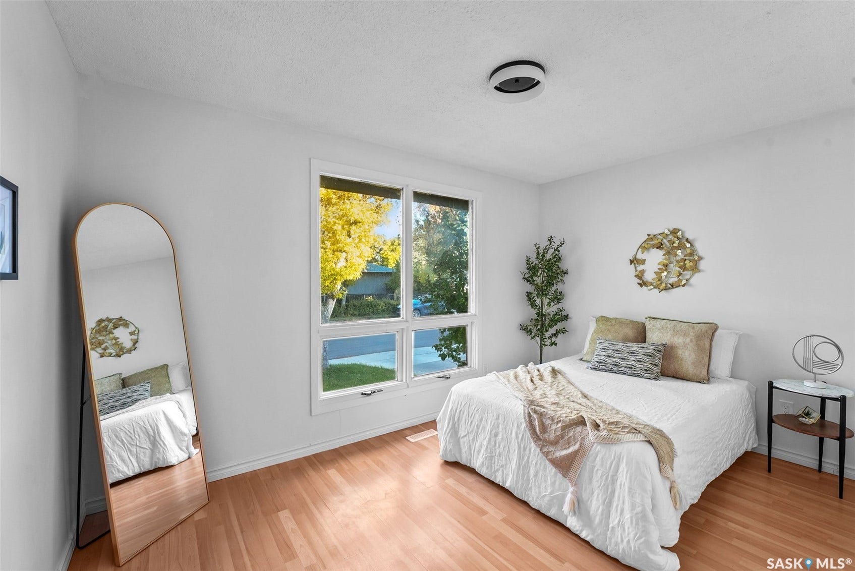 Bright bedroom with wood-finish flooring, large multi-pane windows framing outdoor scenery, an arched mirror, and a nightstand with integrated lighting