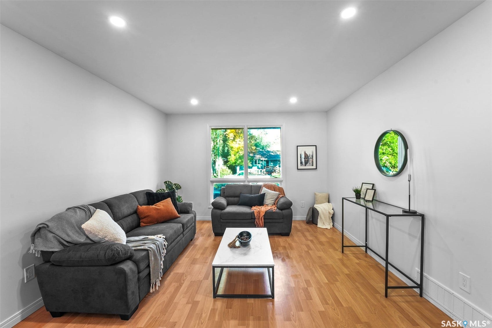 Spacious living area featuring warm wood-finish flooring, recessed ceiling lighting, and multiple seating zones anchored by a white metal-frame coffee table