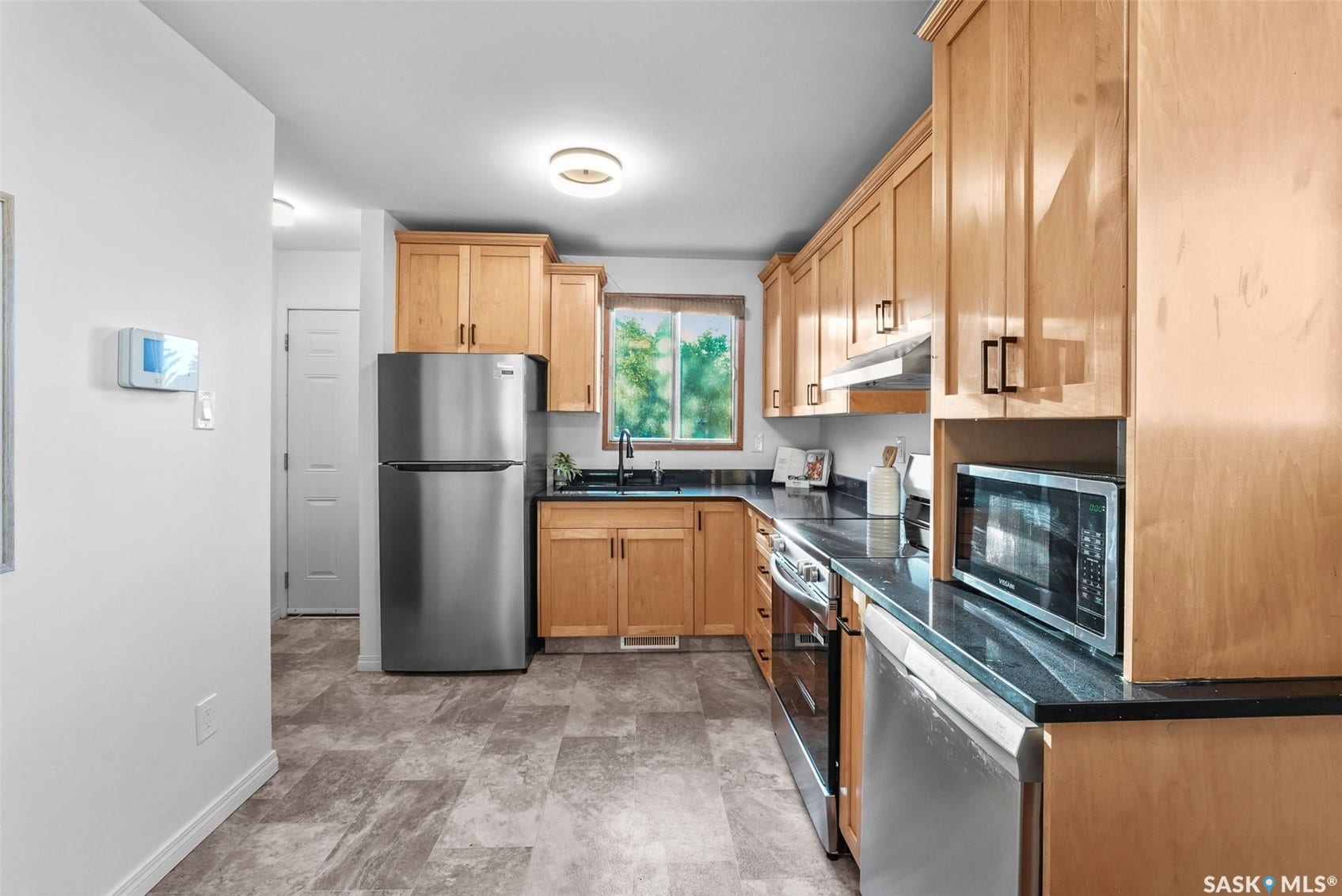 Galley-style kitchen featuring wood-finish cabinetry, stainless steel appliances including a refrigerator and built-in microwave, black countertops, tile backsplash, and a window overlooking landscaping