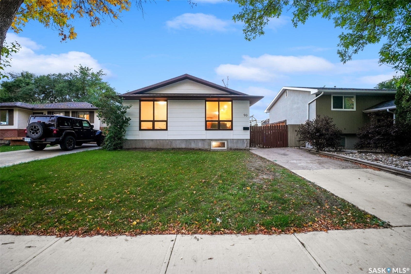 Charming single-story residence featuring a bright, open facade with large illuminated windows, mature landscaping throughout the front grounds, and a spacious manicured lawn with established trees framing the property