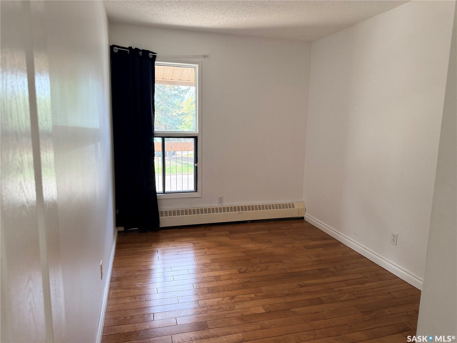 Hardwood flooring, white walls, window with security bars, and a baseboard heating unit