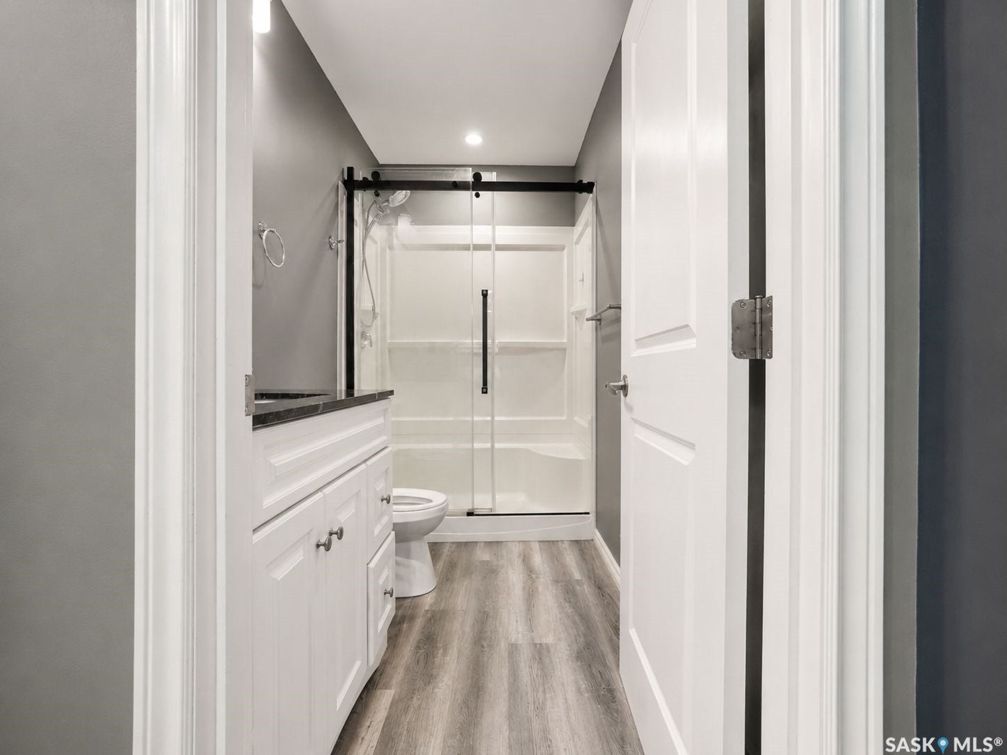 White shaker-style vanity with dark countertop, wood-finish flooring, and a shower with a sliding glass door