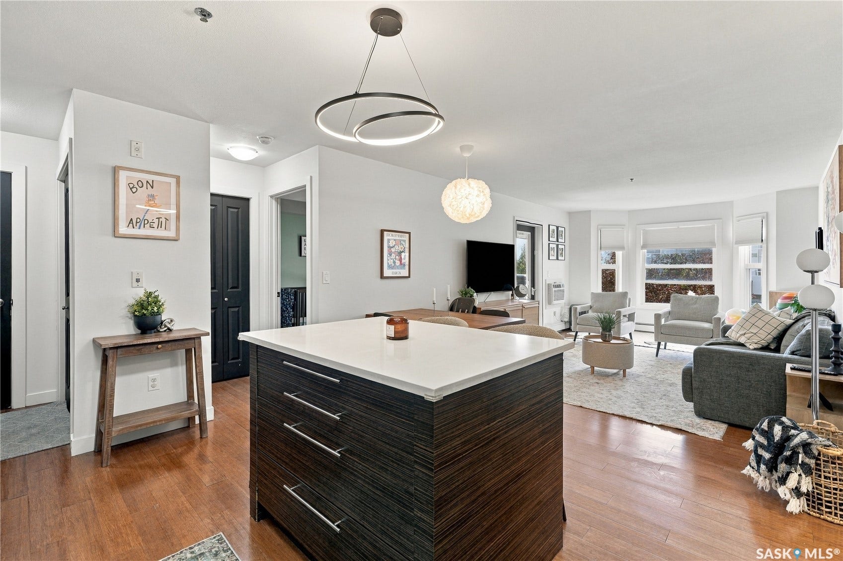 Open-concept living space with wood-finish flooring, a kitchen island with a light-toned countertop, and multiple windows with roller shades