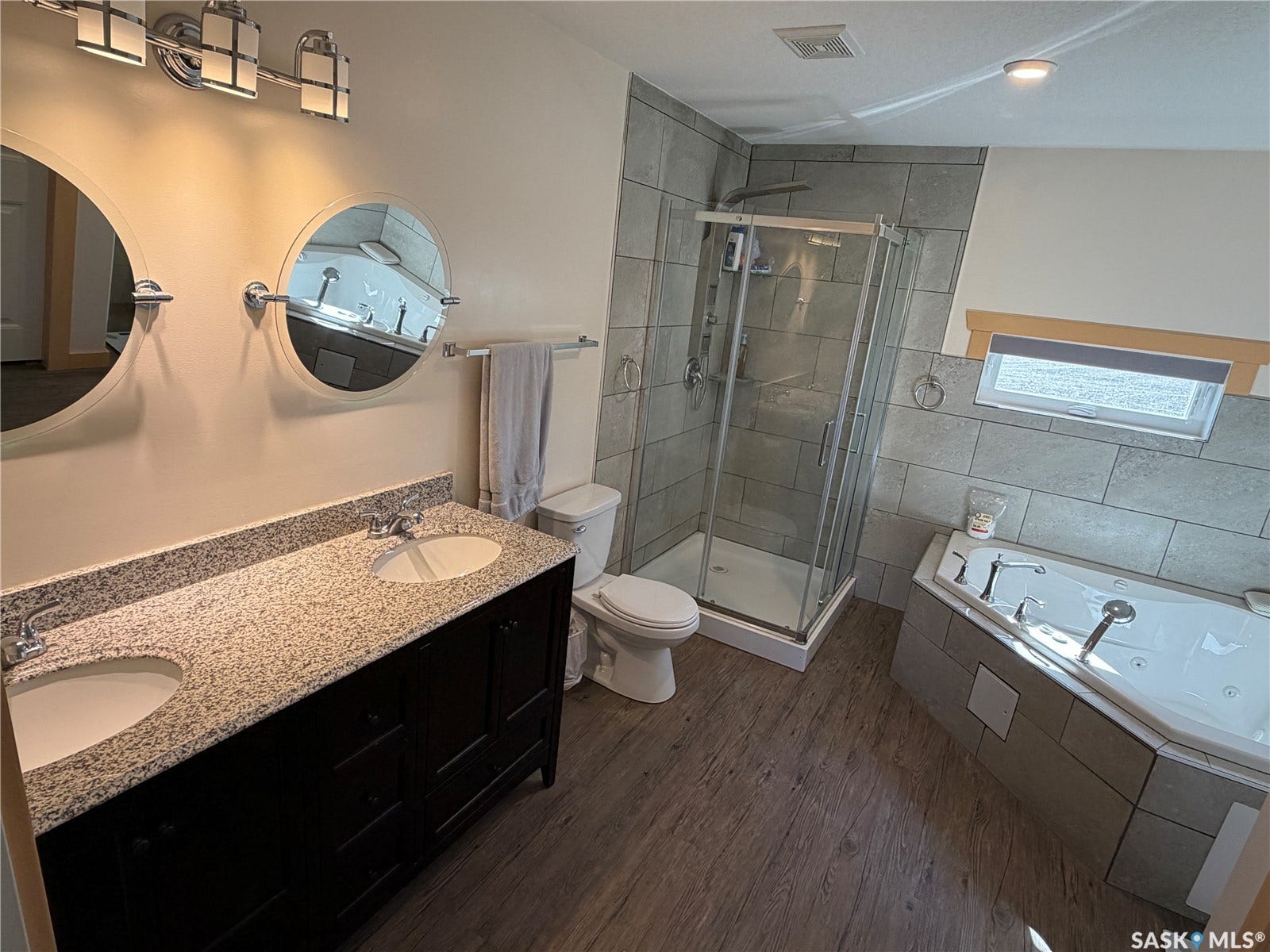 Double vanity with stone countertop, wood-finish flooring, corner jetted tub, and glass-enclosed shower