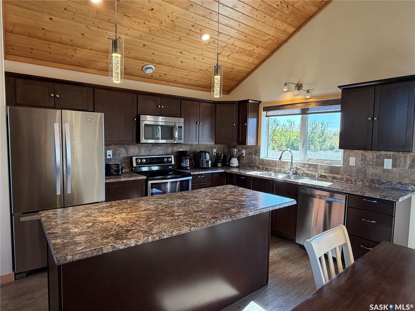 Kitchen featuring vaulted wood-paneled ceilings, stainless steel appliances, dark wood cabinetry, and a central island with a stone-finish countertop