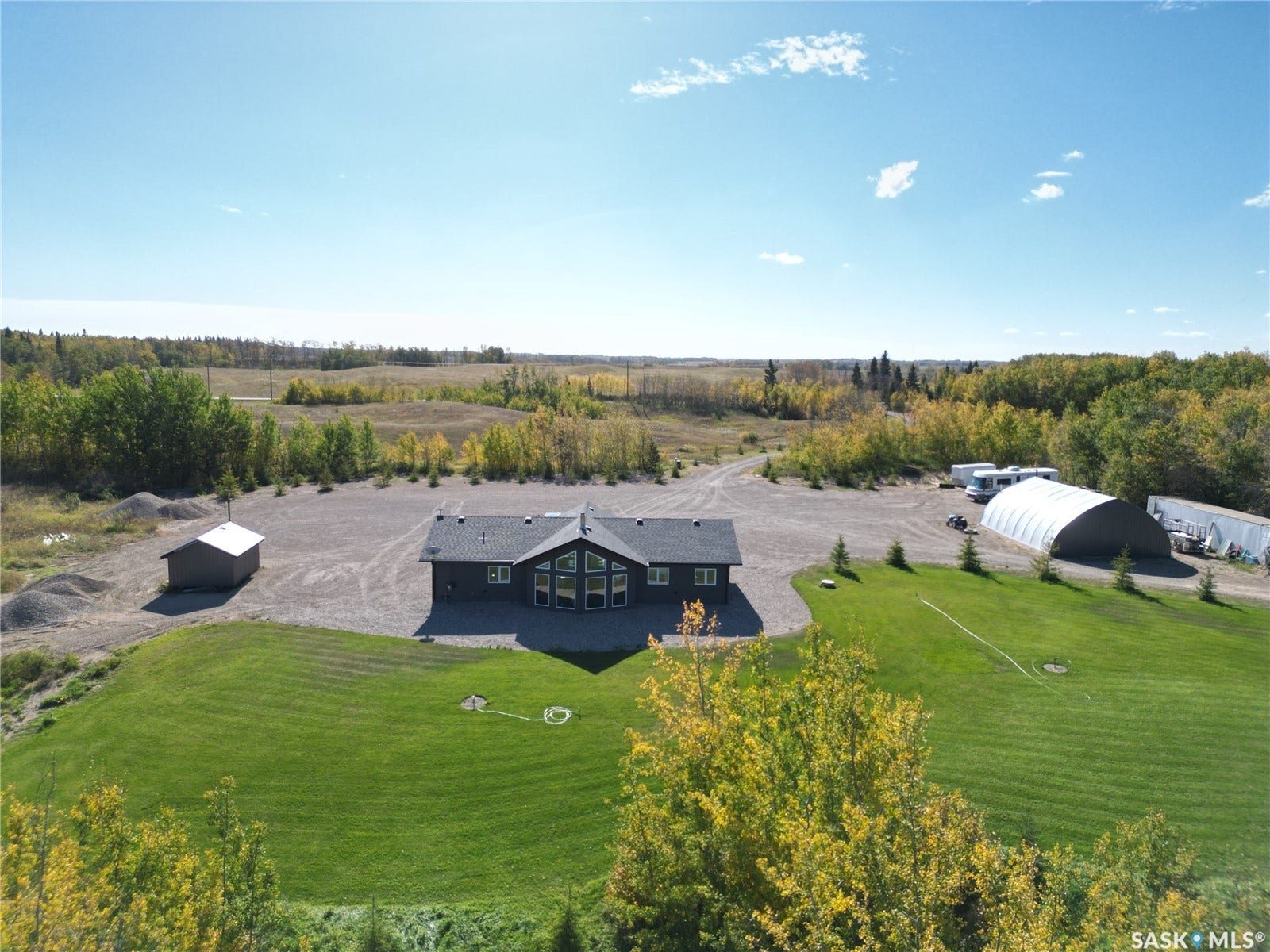 Expansive property featuring a dark-toned residence with multiple large windows, a detached storage shed, a large arch-roofed building, and a vast gravel lot