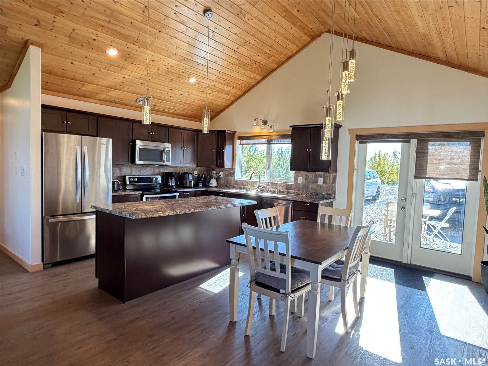 Vance-style ceiling with wood-finish paneling, wood-finish flooring, dark wood-finish cabinetry, stainless steel appliances, and a kitchen island with a stone-finish countertop