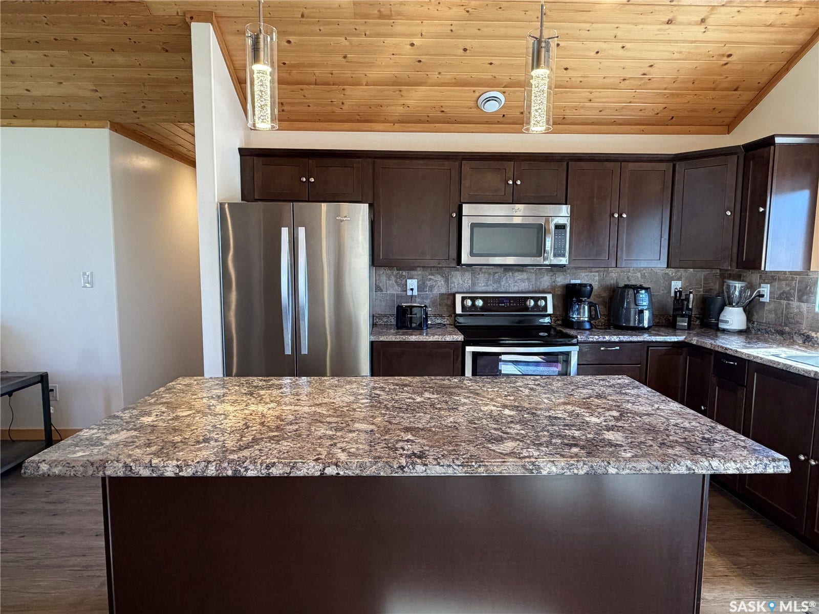 Wood-paneled vaulted ceiling, dark wood-finish cabinetry, stainless steel appliances, speckled countertops, and a kitchen island