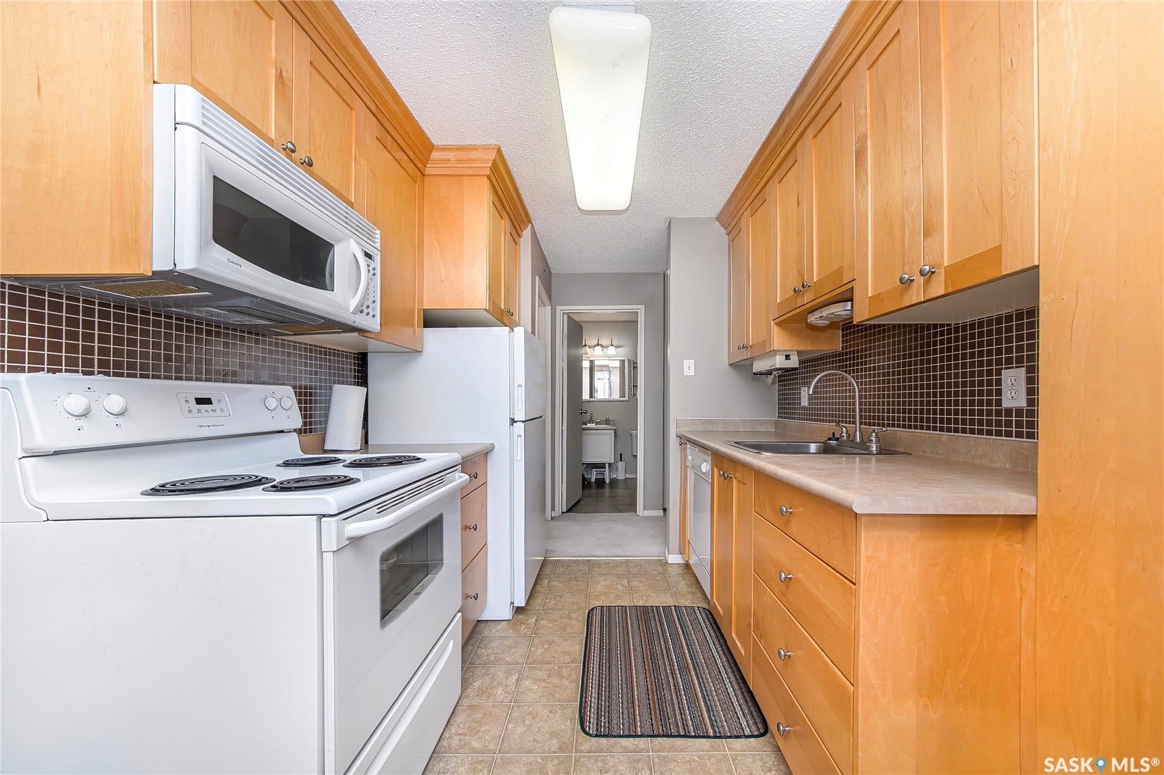 Galley kitchen featuring light wood-finish cabinetry, a ceramic tile backsplash, and a white appliance package