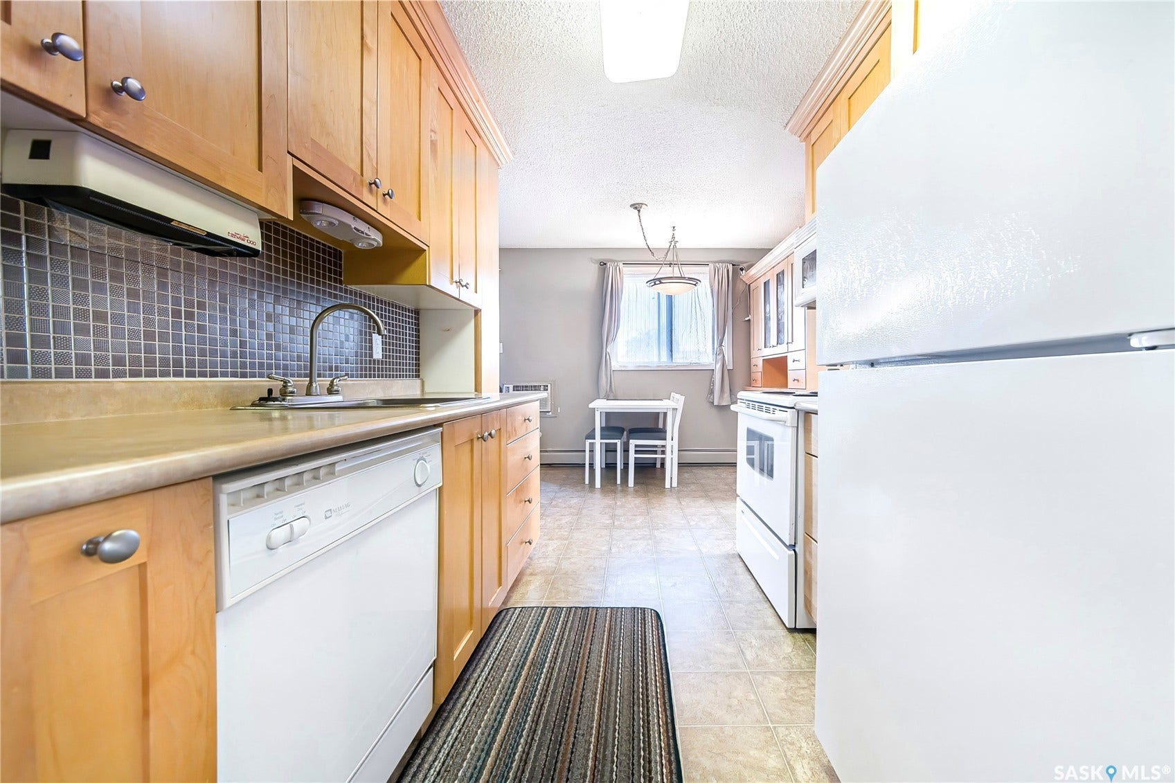 Light wood-finish cabinetry and a mosaic tile backsplash highlight this kitchen, featuring white appliances, a stainless steel sink, and durable tile flooring