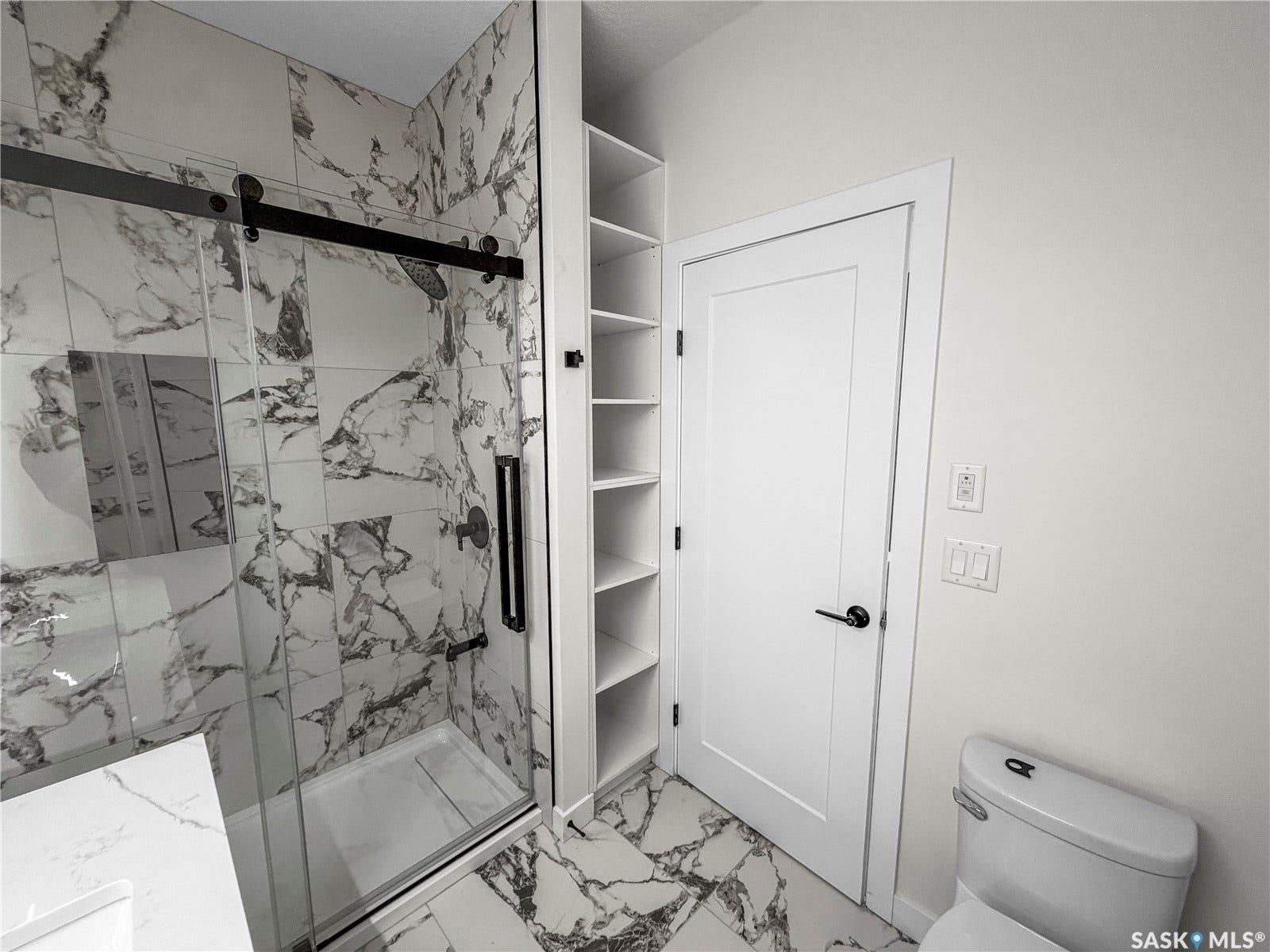 Walk-in shower with a sliding glass door and matte black hardware, floor-to-ceiling marbled tile, and built-in shelving