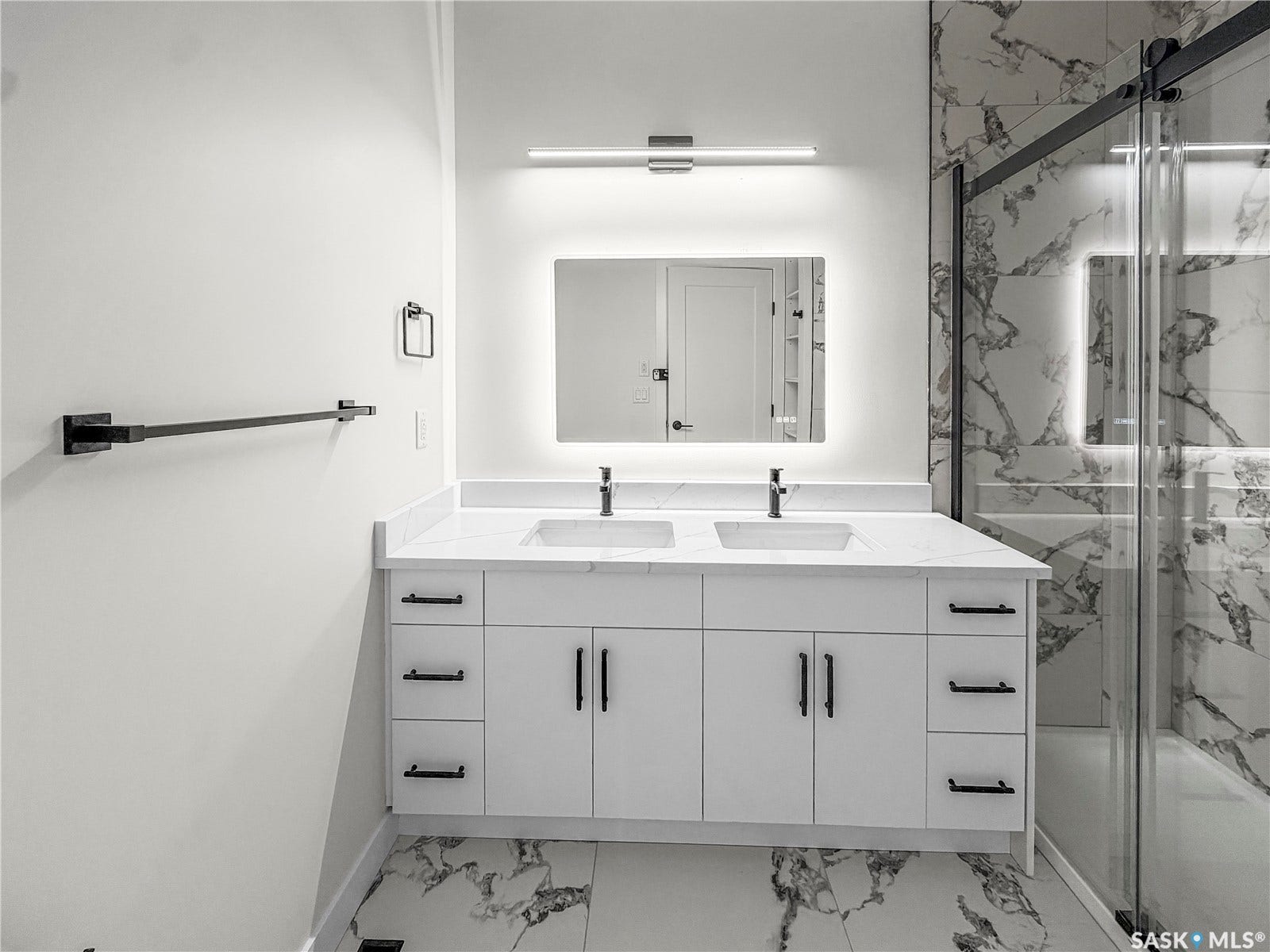 Dual vanity with integrated sinks, white cabinetry with matte black hardware, back-lit mirror, and full-height marble-look tile