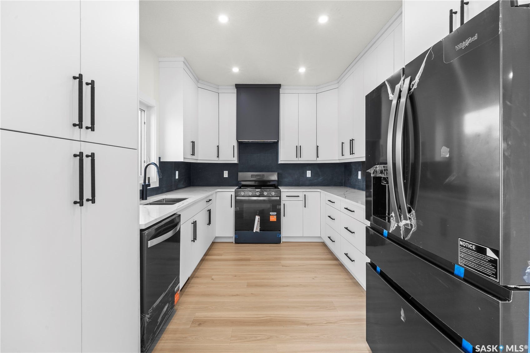 White cabinetry with black hardware, U-shaped kitchen layout, dark backsplash, stainless steel appliances, and wood-finish flooring