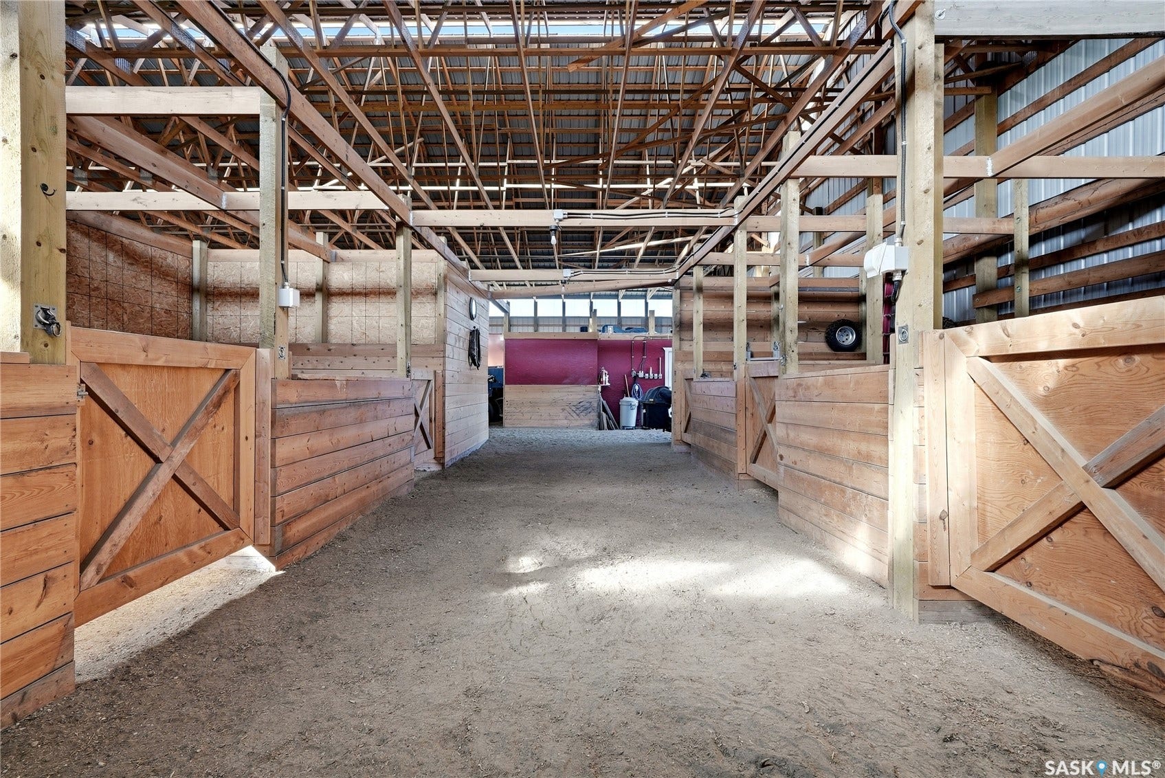 Barn interior featuring exposed wood beam construction, wood-paneled stalls with cross-brace doors, and a sand floor
