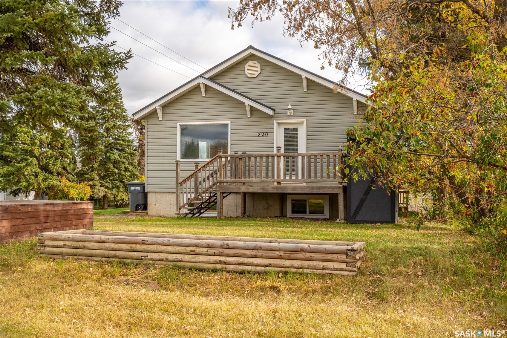 Gray horizontal siding exterior with white trim, front deck with wood railing, white front door, and a visible basement window