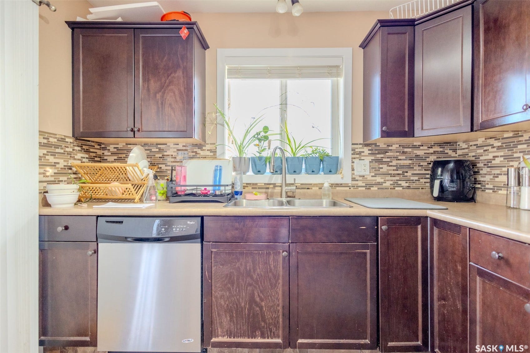 Kitchen featuring dark wood-finish cabinetry, tile backsplash, stainless steel dishwasher, and a double basin sink with a window overhead