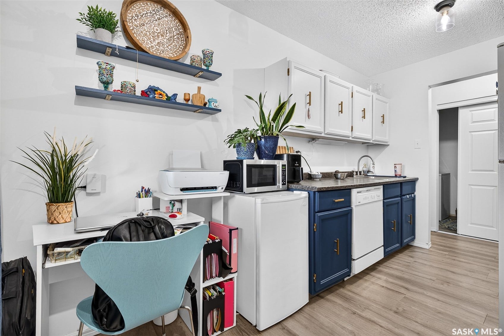 White upper cabinetry with gold-tone hardware, dark blue lower cabinetry, light wood-finish flooring, and a compact desk area with open shelving