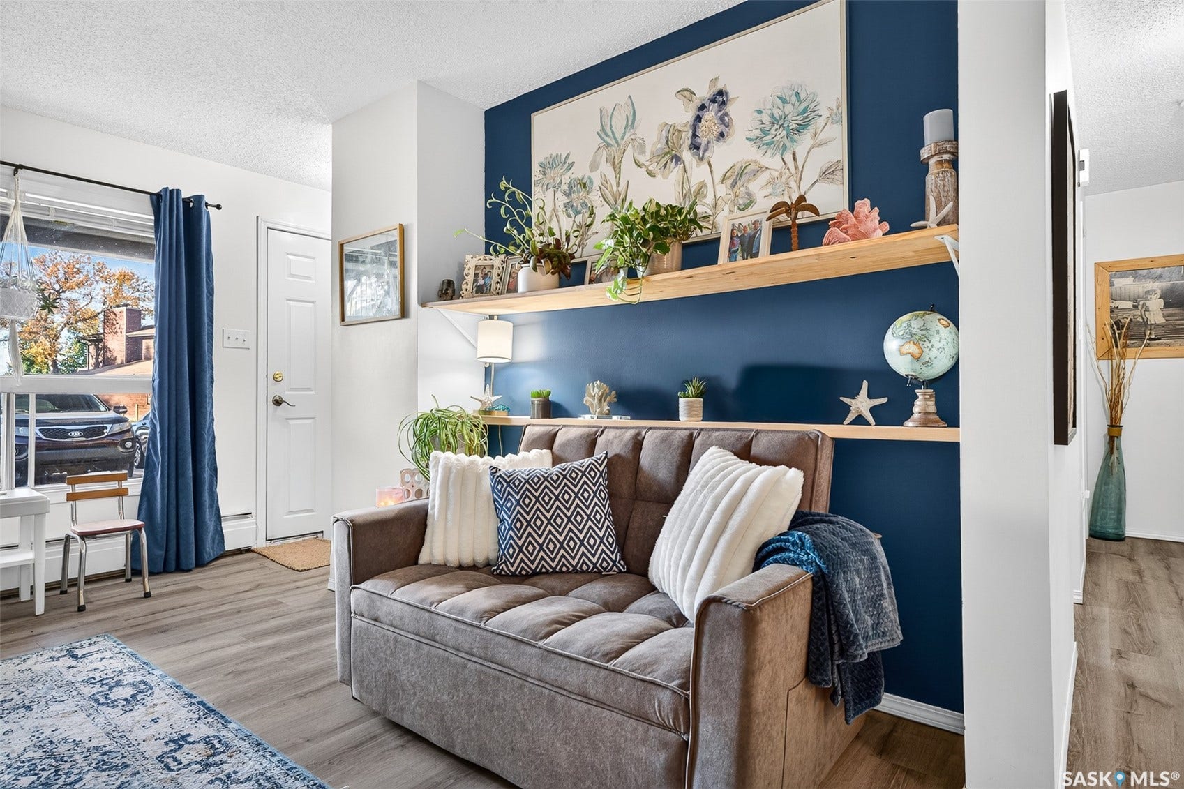 Stylish living room featuring a deep navy accent wall with two wood-finish floating shelves, warm wood-finish flooring, and a large window with blue drapery